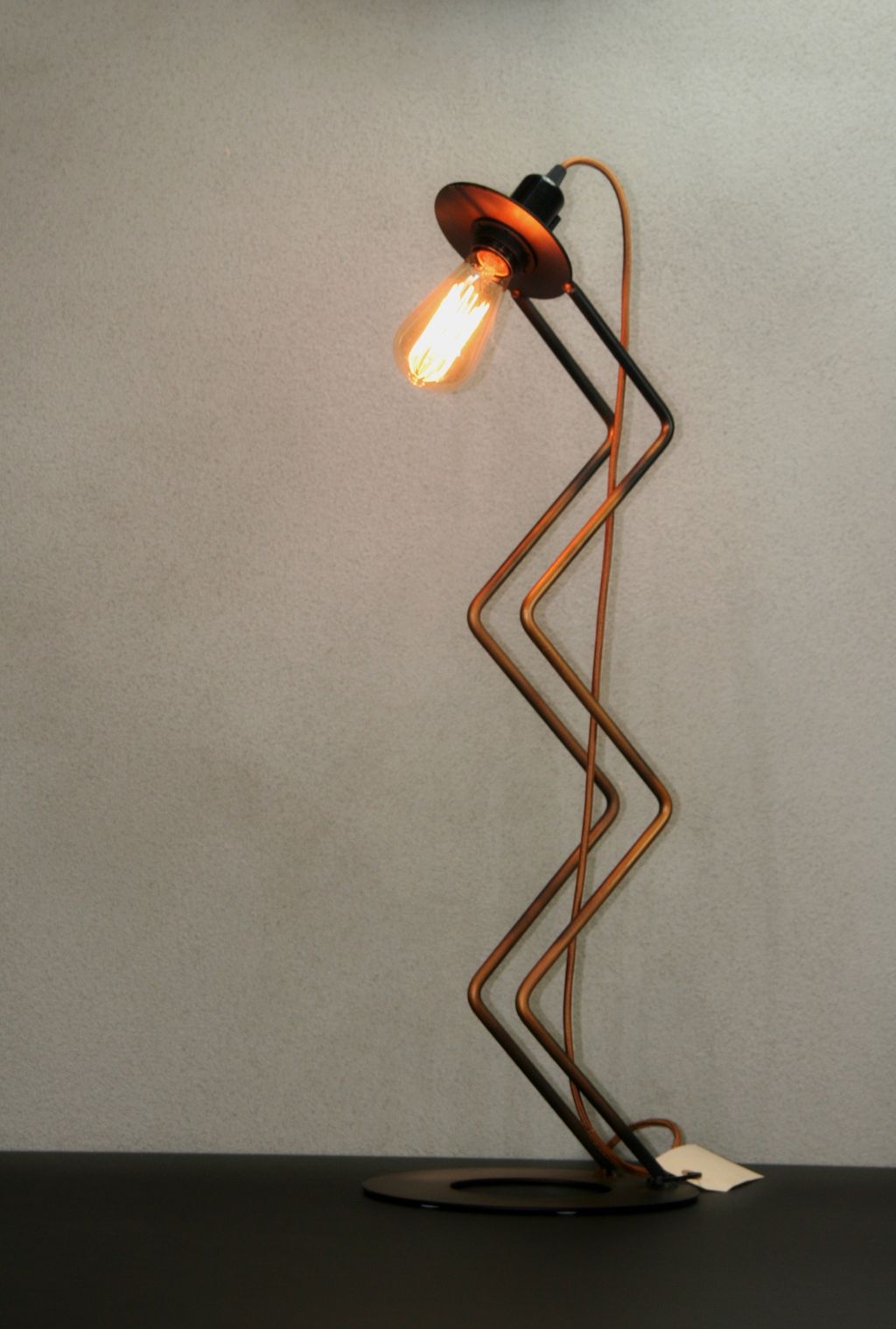 Høj zig zag lampe i bronze farver, står tændt på et bord